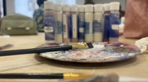Close-up view of a paint-stained artist’s palette with brushes resting on it, set on a wooden table. In the background, several bottles of paint are lined up, slightly out of focus, along with bags and blurred figures of people in a creative studio or classroom setting.